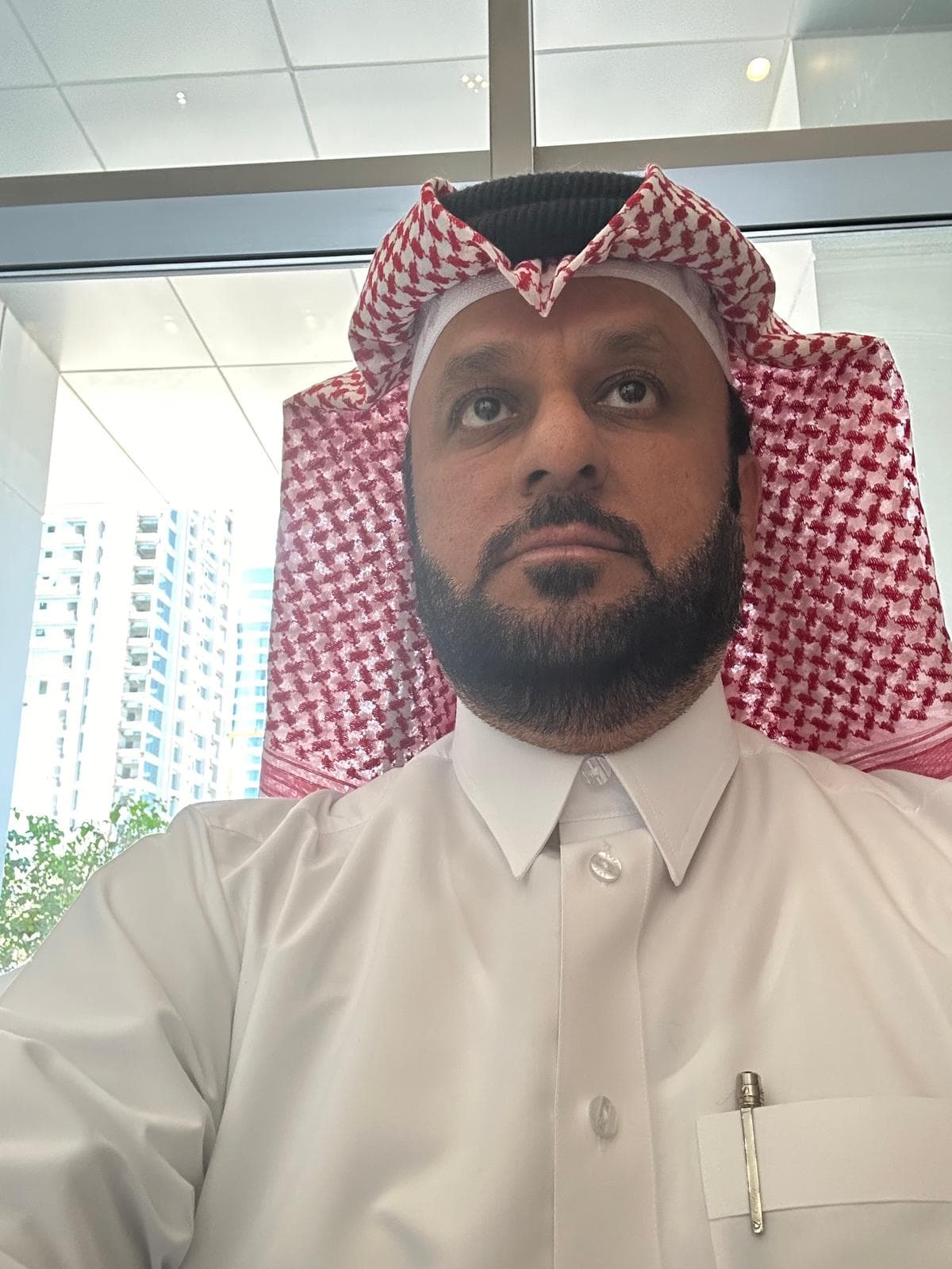 Bearded man wearing a white thobe and red and white checkered ghutra, looking upwards.