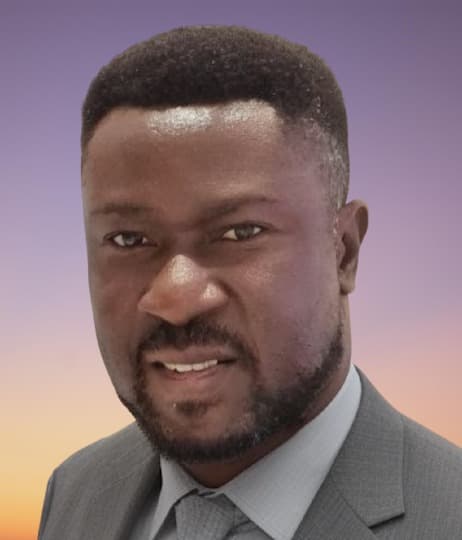 Smiling Black man with a beard in a grey suit against a sunset gradient background.