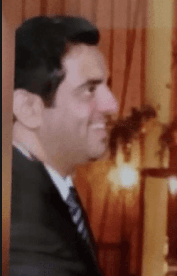 Smiling man in profile wearing a dark suit and striped tie against a warm background.